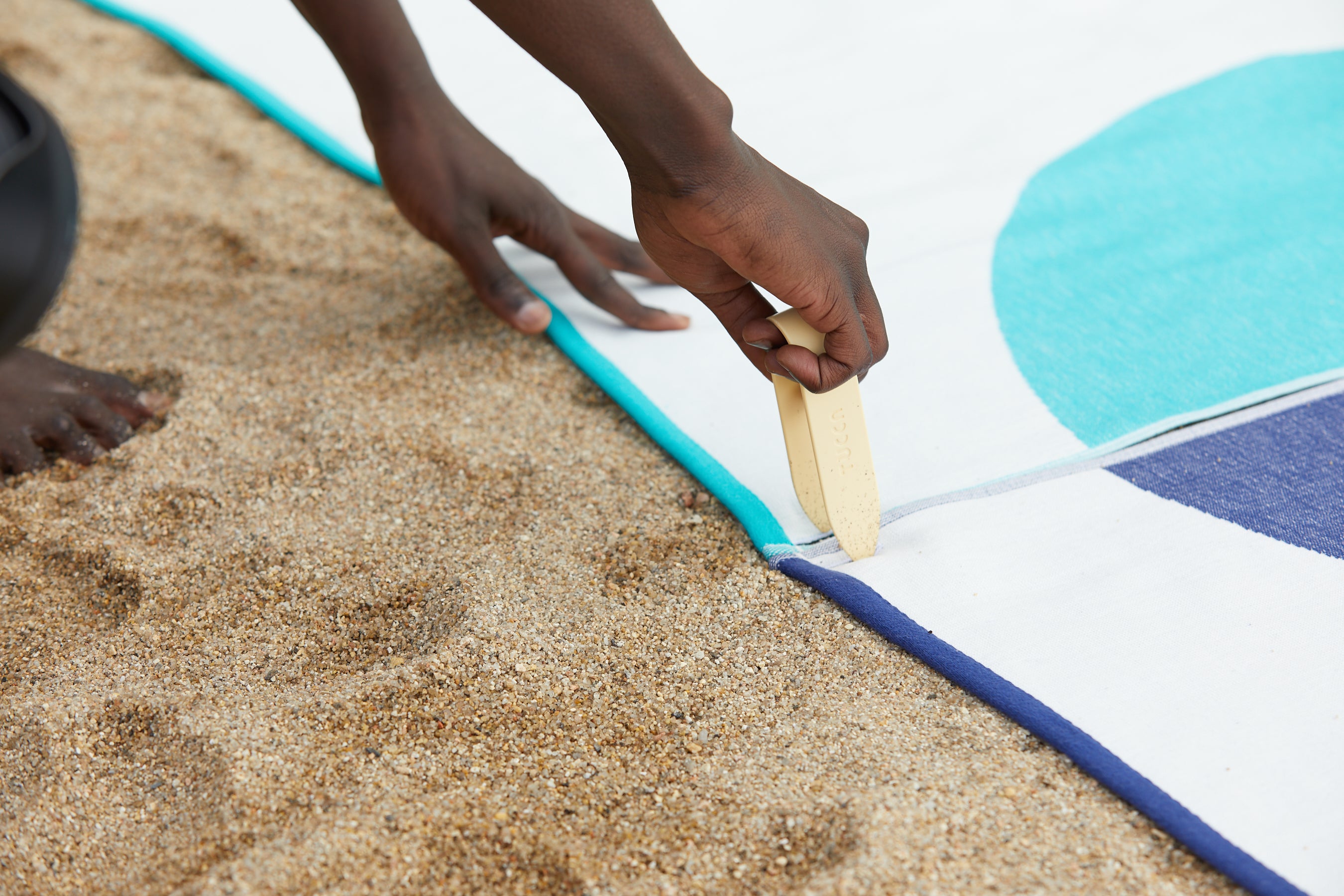 Picture showing how to fix your beach towels into the sand so they don't get blown away by the wind. Inserting the Tucca pin through the corners of the premium light beach towels Florida and Mayeri styles. This way both beach towels can be connected 