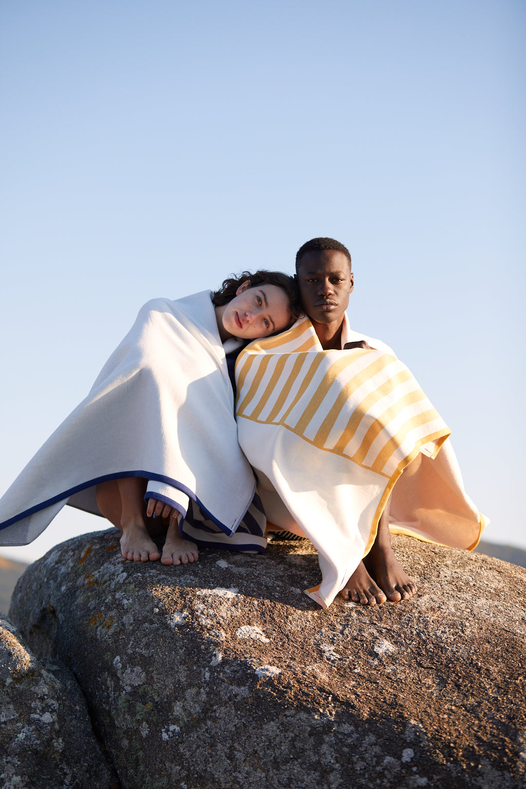 Girl and boy covering themselves with the Tucca Florida and Danai beach towels styles. Sitting on a rock in the beach. Holding both Tucca towels, beach towels that doesn't get blown by the wind. Super soft texture as it is made with 100% organic cotton.
