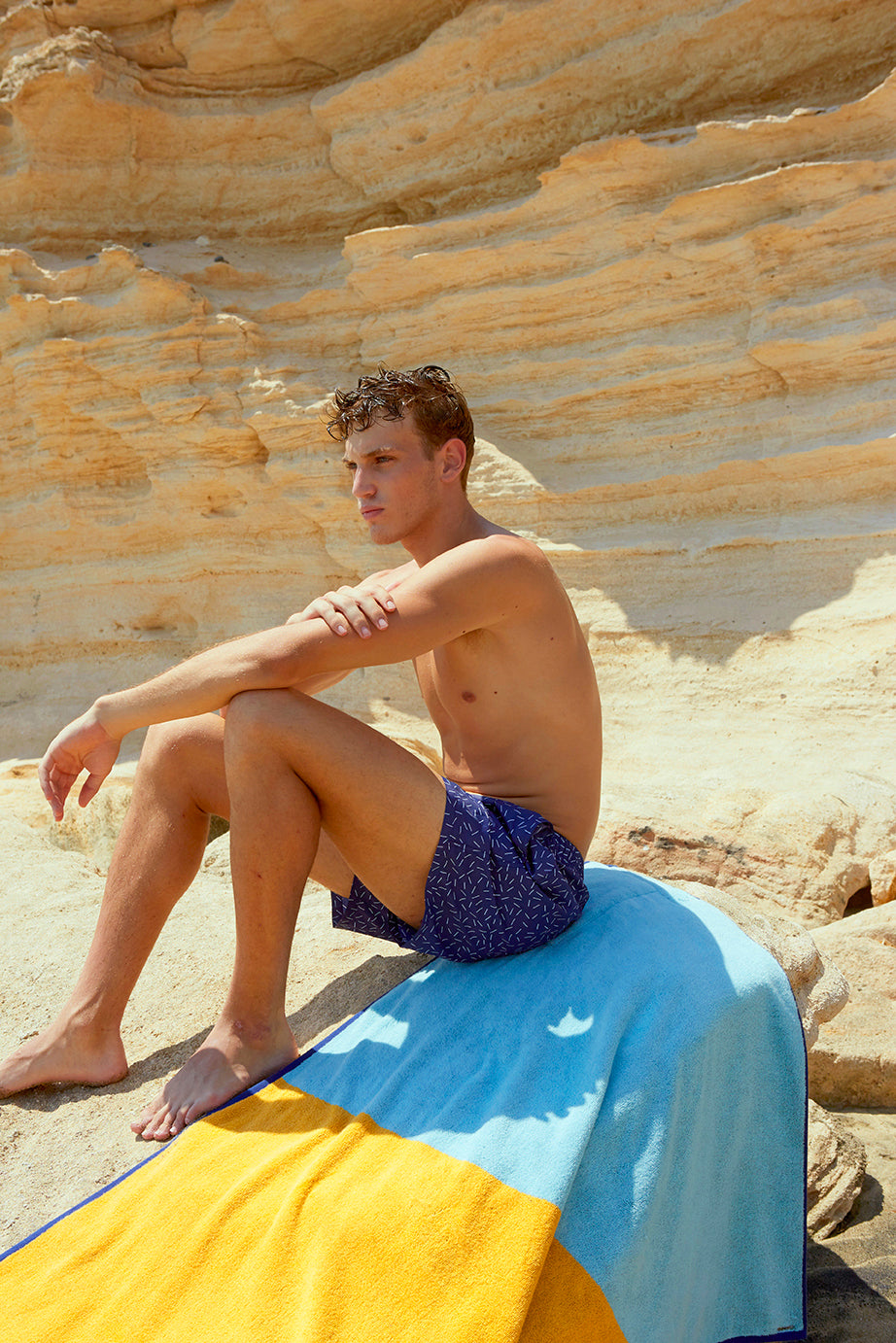 Man set on a rock over his Dune Tucca beach towel extended. Tucca "Dune" beach towel extended. Light blue, camel yellow and dark blue colors in big blocks composing a beautiful design. Large beach towel that doesn't get blown by the wind. Thick and soft texture as it is made with 100% cotton.