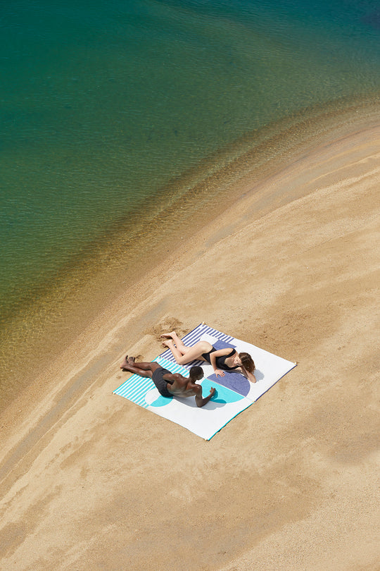 Two beach towels connected without sand in between and creating a bigger common space in the beach. Couple enjoying the sun in the beach lying on the Florida and Mayeri styles, premium beach towels, extra light and made of 100% organic cotton.